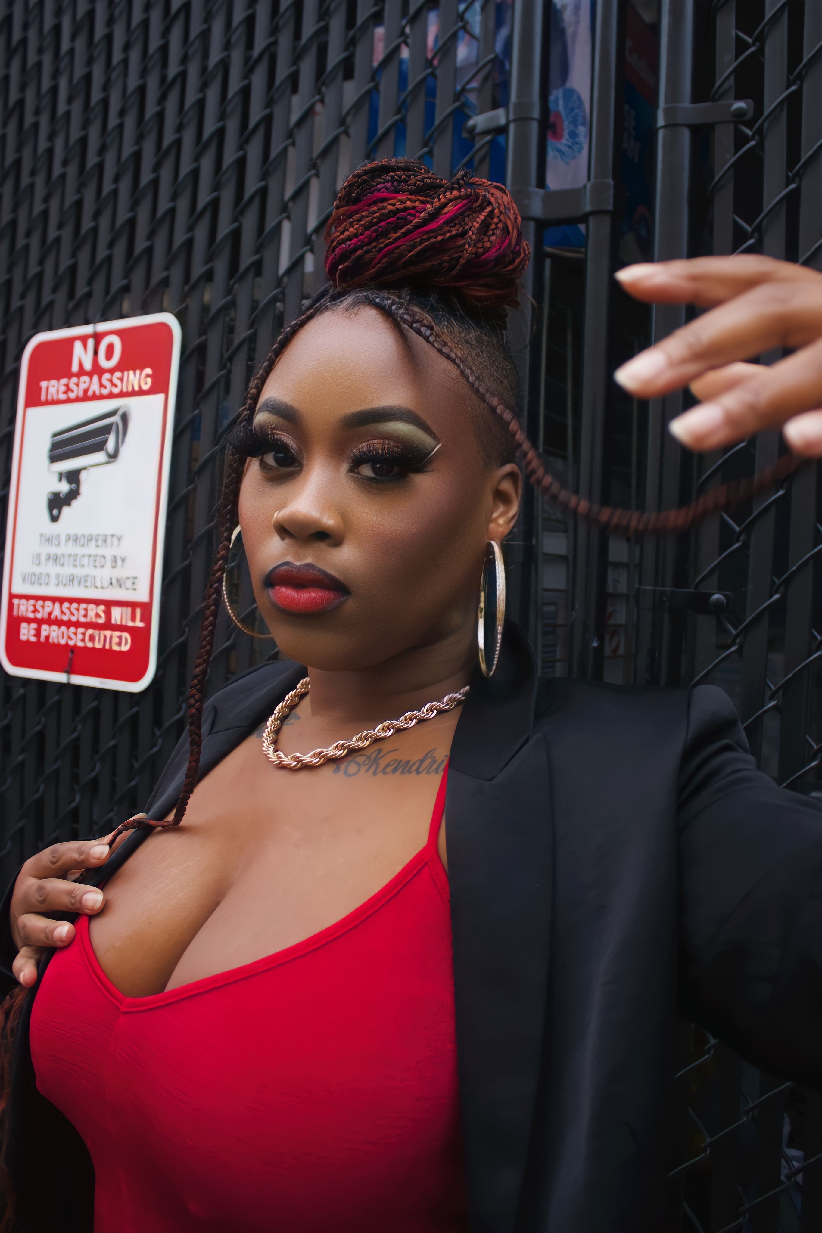 A stylish woman in a red dress and black coat poses confidently by a metal gate with bold signage.