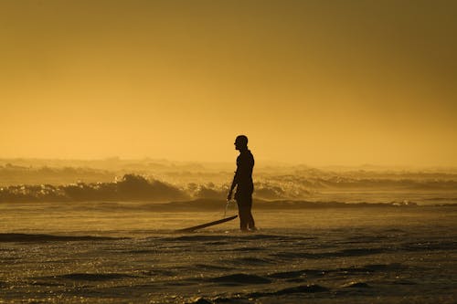 일몰 동안 자신의 서핑 보드를 들고 해변에 서있는 남자