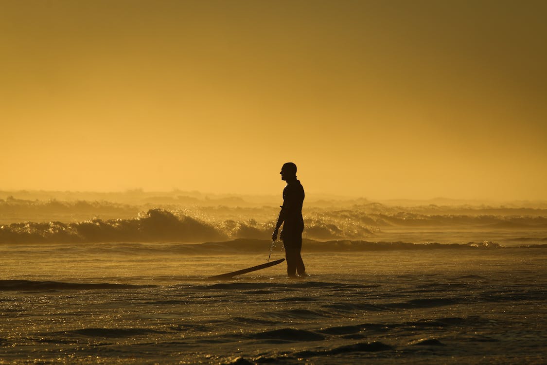 Gratis Pria Berdiri Di Tepi Laut Sambil Memegang Papan Selancar Saat Matahari Terbenam Foto Stok