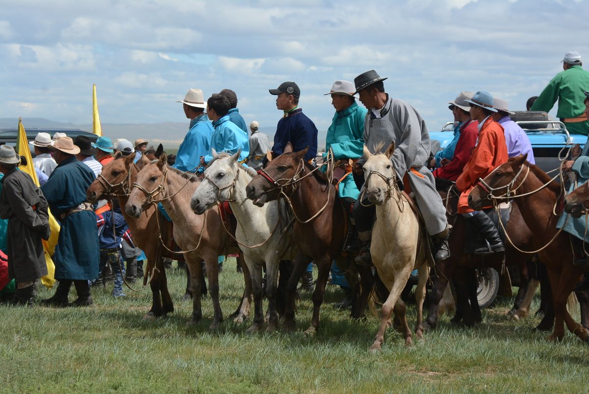 Riders in colourful deel coats and wide-brimmed hats lined up on horseback before a race, some holding yellow flags, open steppe behind them