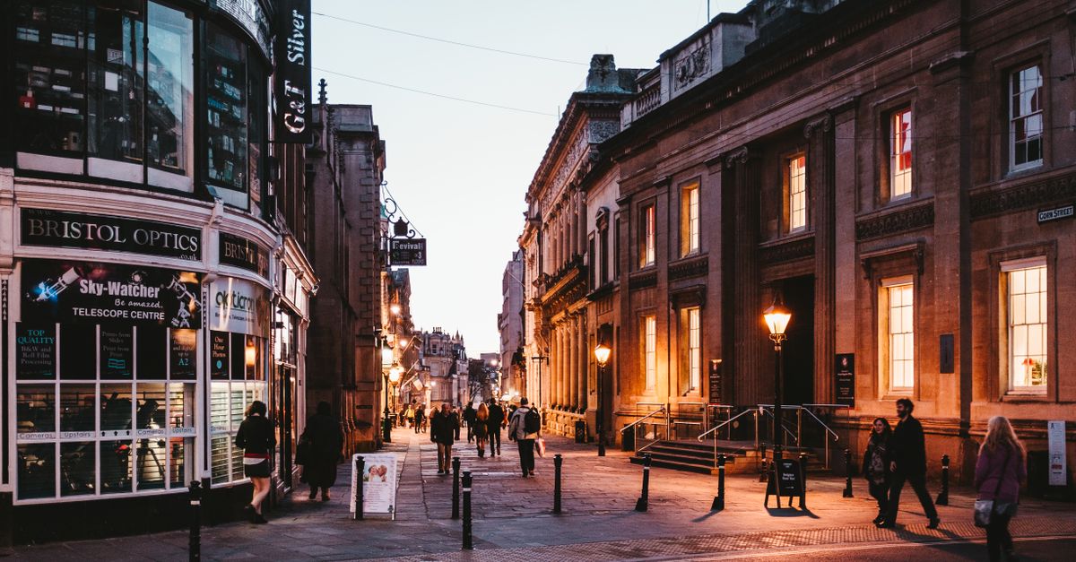 People walking along illuminated street in Bristol, England, during twilight with historic buildings.