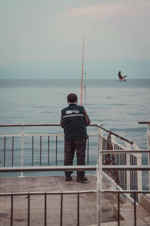 Free An adult fisherman stands with a fishing rod on a lakeside dock under a cloudy sky, enjoying a peaceful leisure activity. Stock Photo