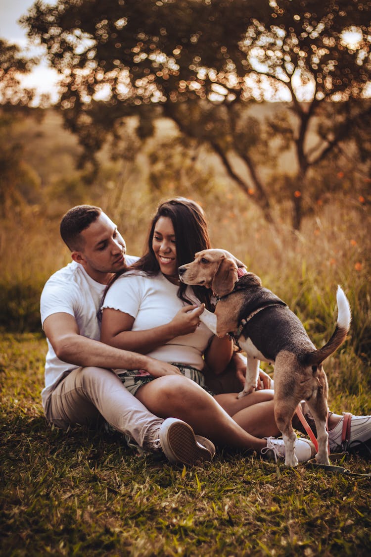 Happy Couple With Dog Sitting On Ground In Field