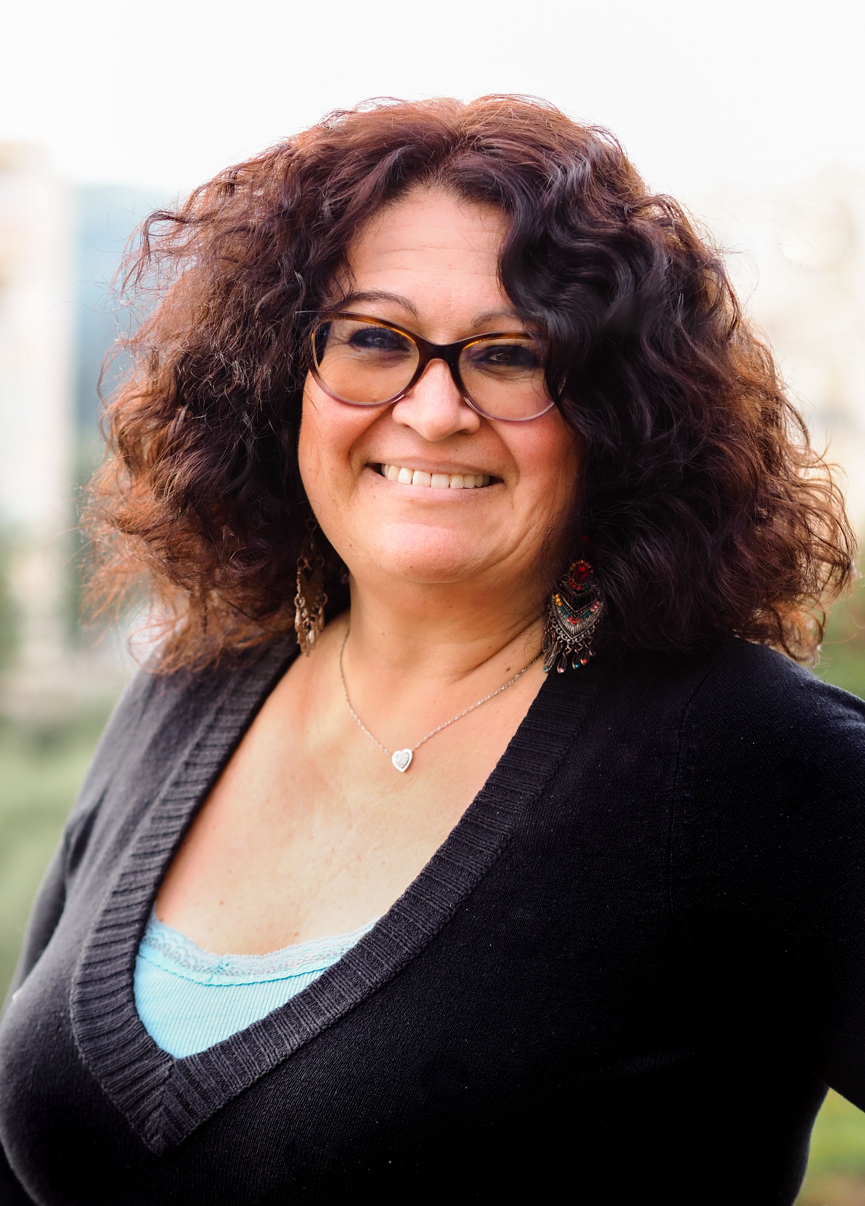 Portrait of a woman with curly hair and glasses smiling warmly in an outdoor setting.