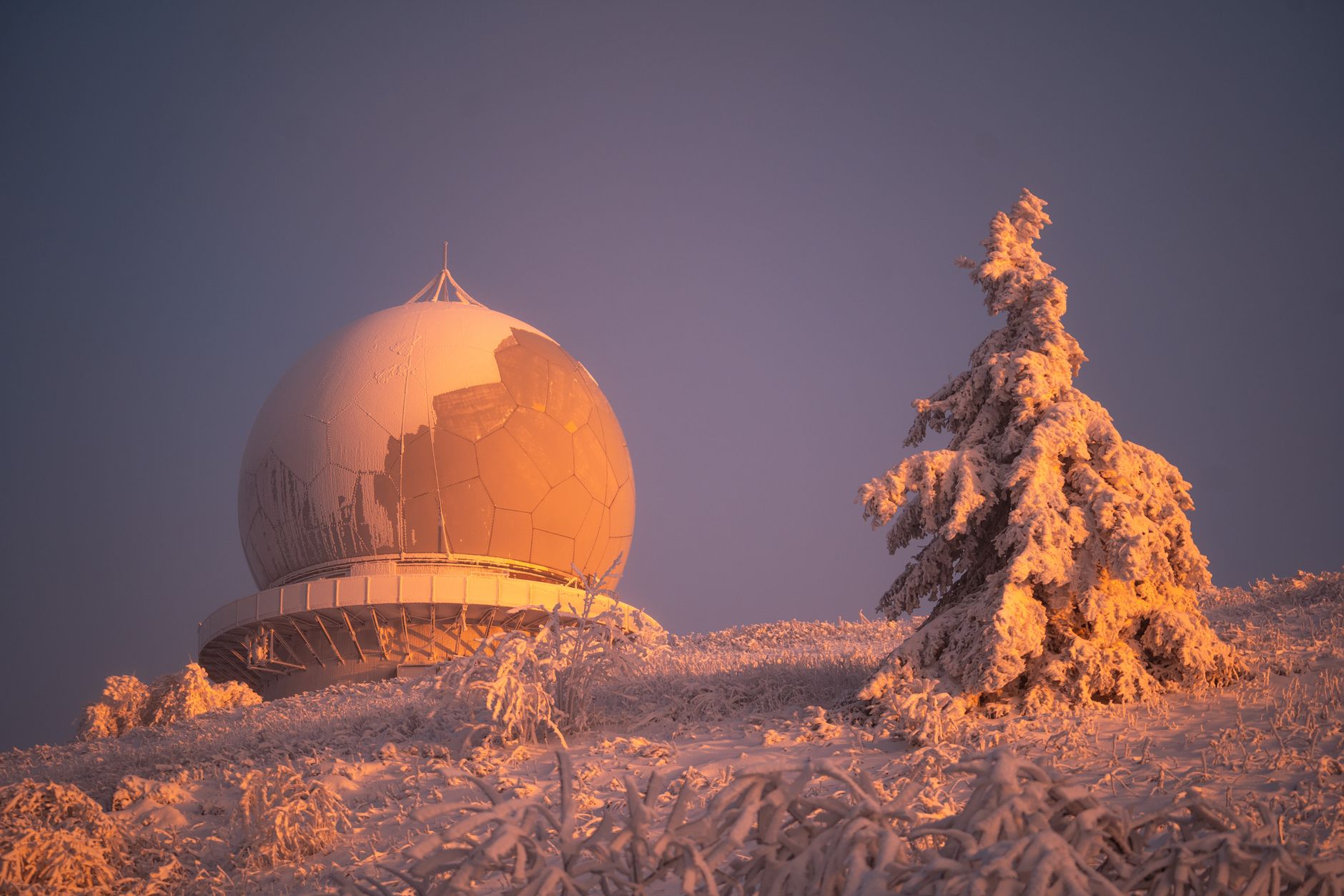 Serene winter landscape at sunset featuring a frosted radar dome and snow-covered trees.