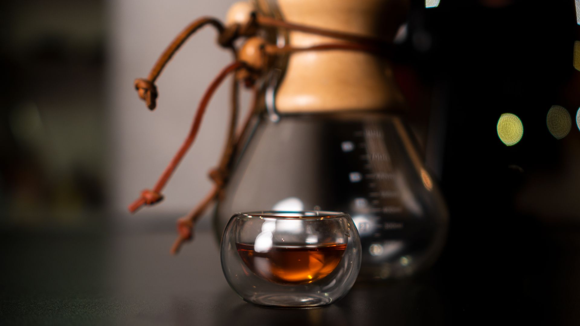 Close-up of a double-walled glass cup with coffee against a blurred Chemex coffee maker backdrop.