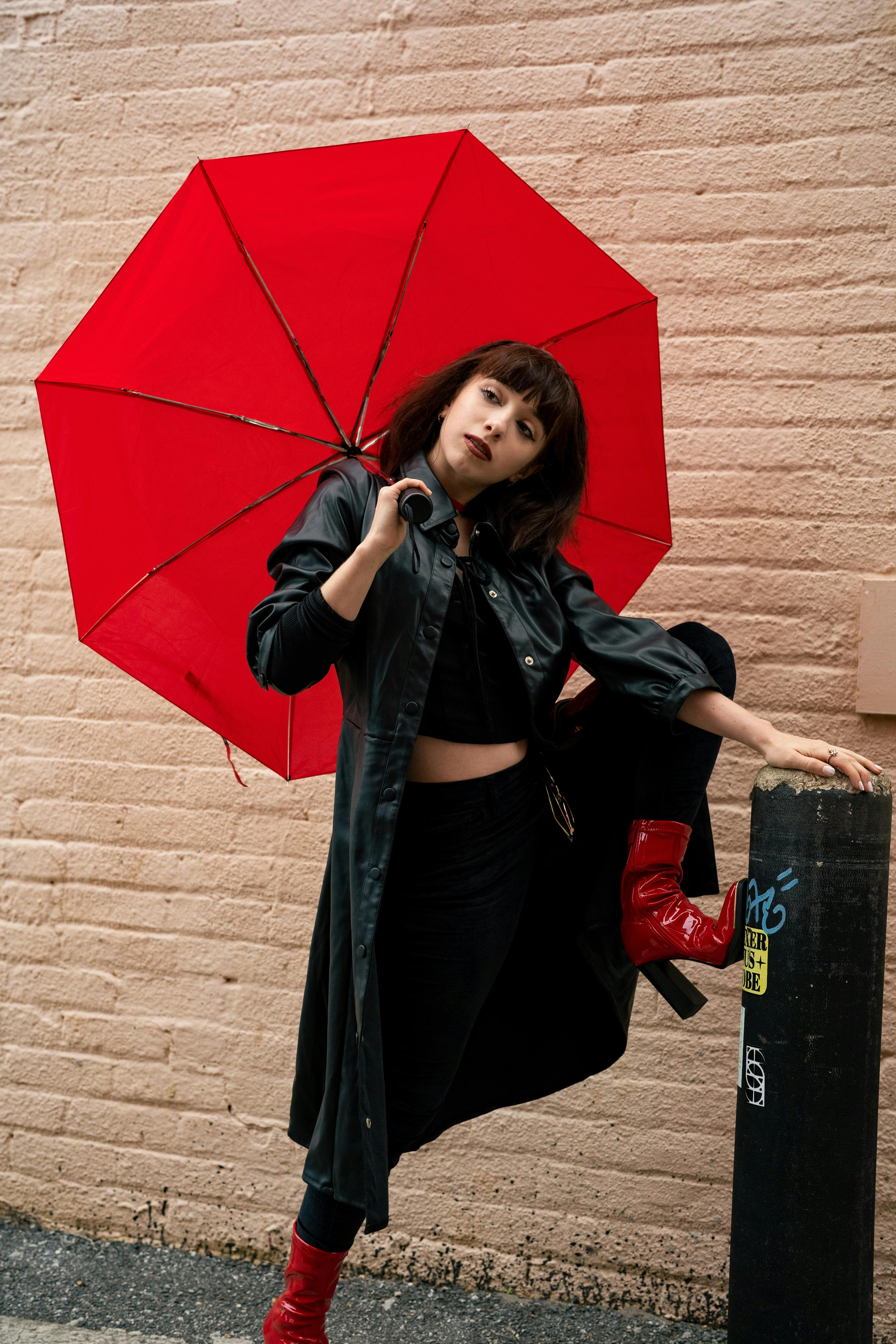 Free Stylish woman in black coat and red boots poses with a red umbrella against a brick wall. Stock Photo