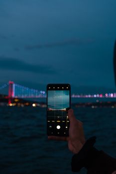 Silhouette of a hand holding a smartphone photographing a lit-up bridge at night.