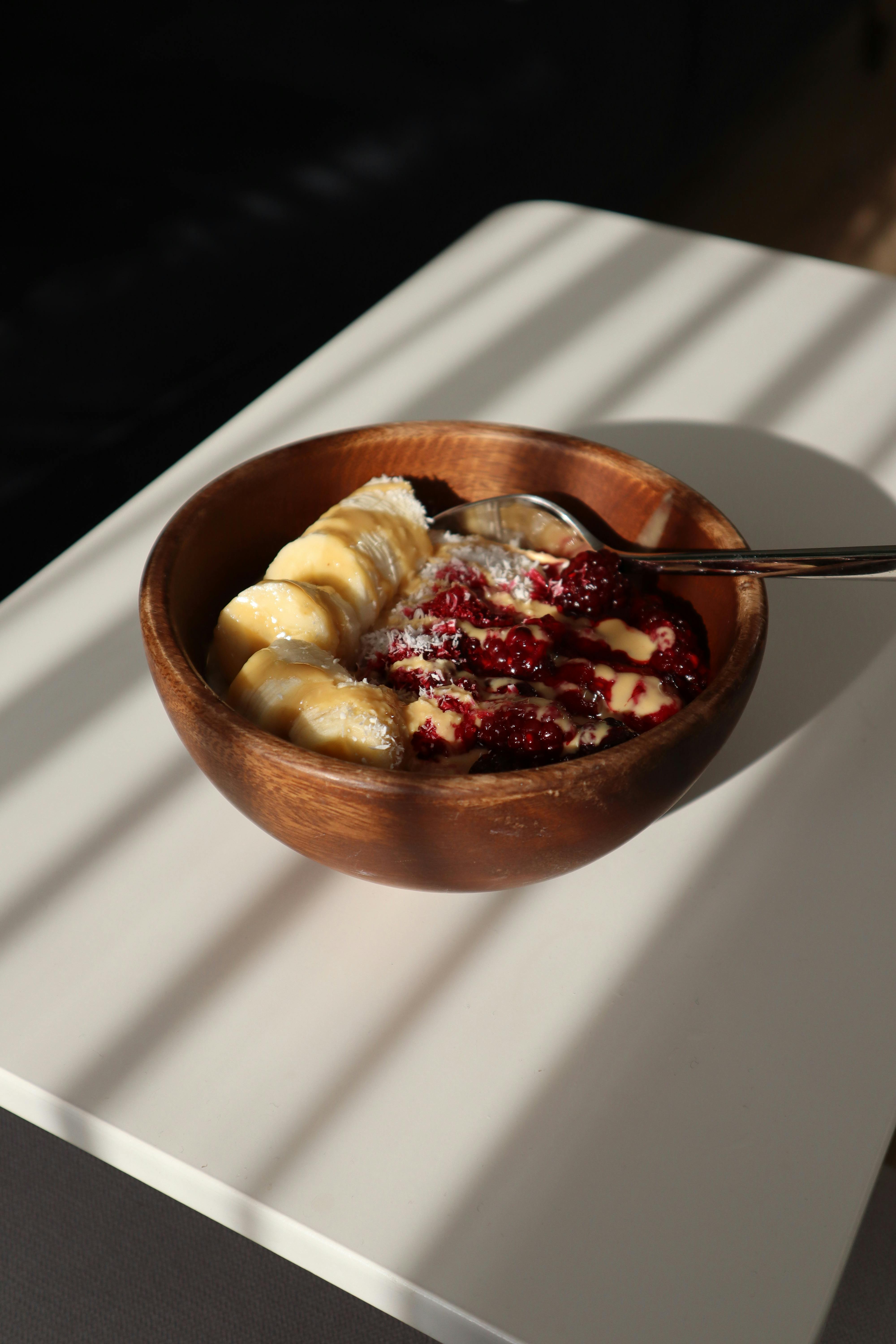 A wooden bowl of fresh bananas and raspberries with yogurt on a table.