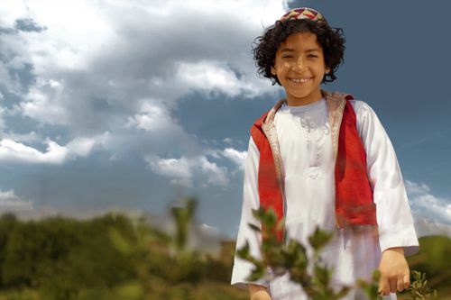 Free A cheerful young boy with curly hair wearing traditional clothing, smiling against a scenic nature backdrop. Stock Photo