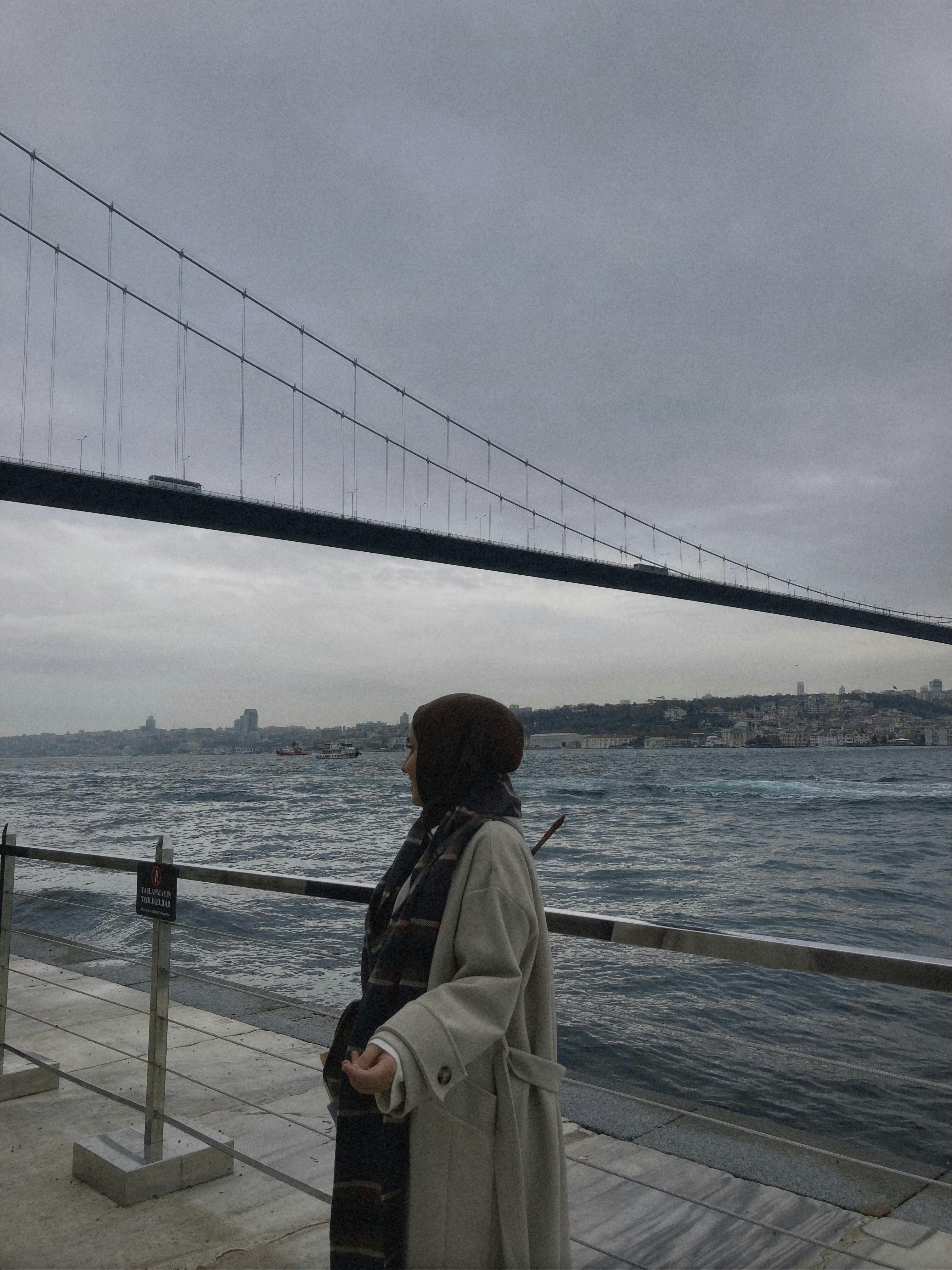 A woman in a trench coat stands by the Bosphorus with the bridge in view on a cloudy Istanbul day.