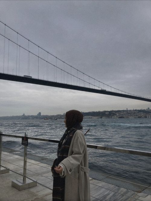Free A woman in a trench coat stands by the Bosphorus with the bridge in view on a cloudy Istanbul day. Stock Photo