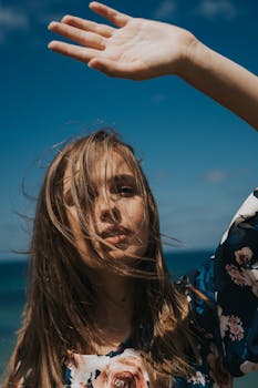 A woman with long hair poses with hand raised against a clear blue sky, capturing a moment of serene beauty.