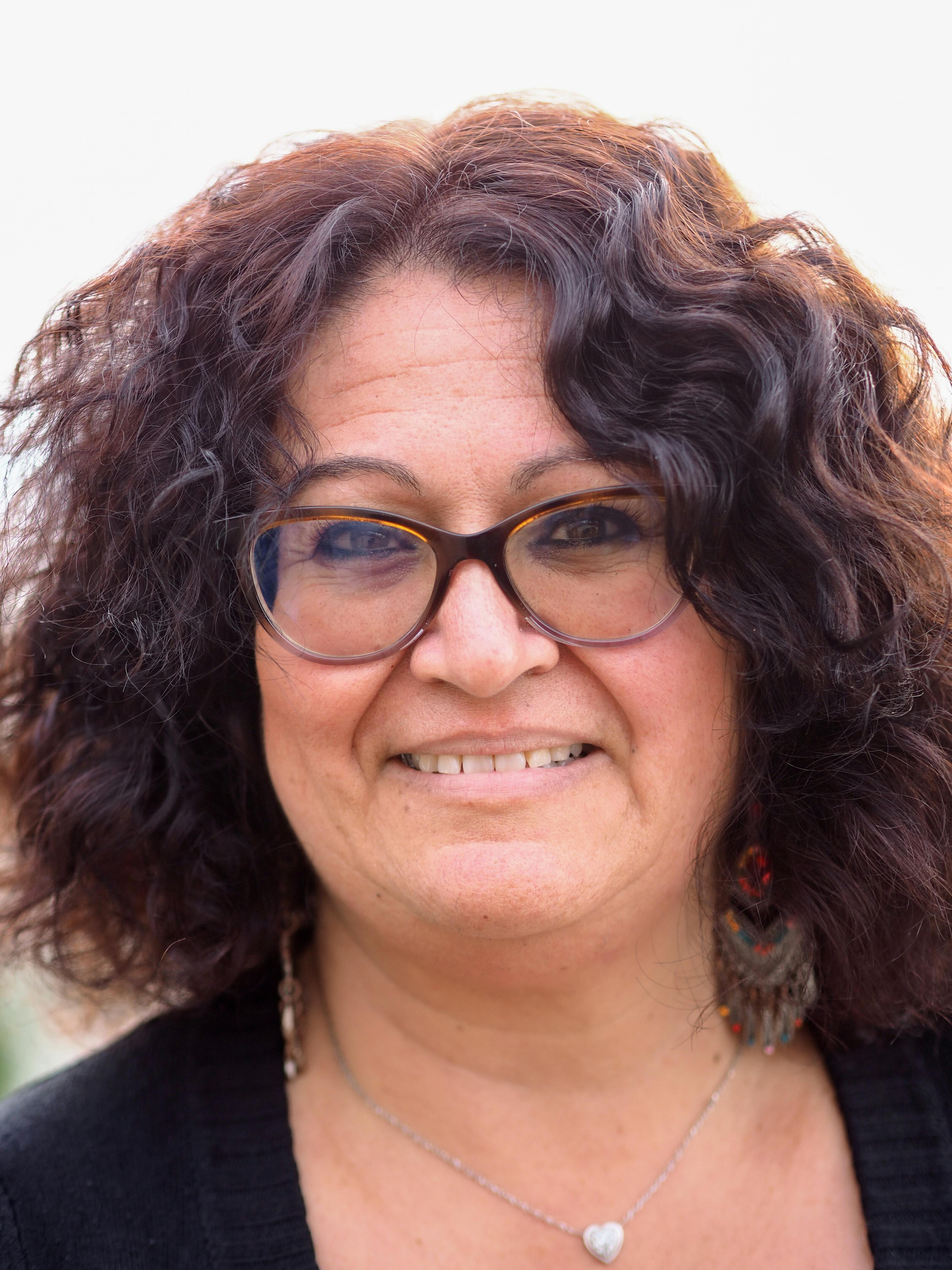 Portrait of a cheerful senior woman with curly brunette hair, wearing glasses and a necklace, smiling warmly.