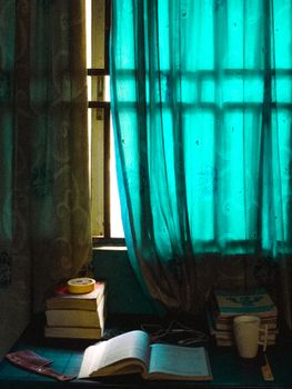 A peaceful reading corner with books, a mug, and sunlight filtering through turquoise curtains.