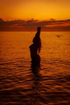 A woman raises her arms in silhouette against a vivid orange sunset over the ocean waves.