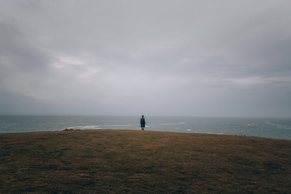 A lone person stands on a grassy cliff, overlooking a stormy sea with moody clouds.