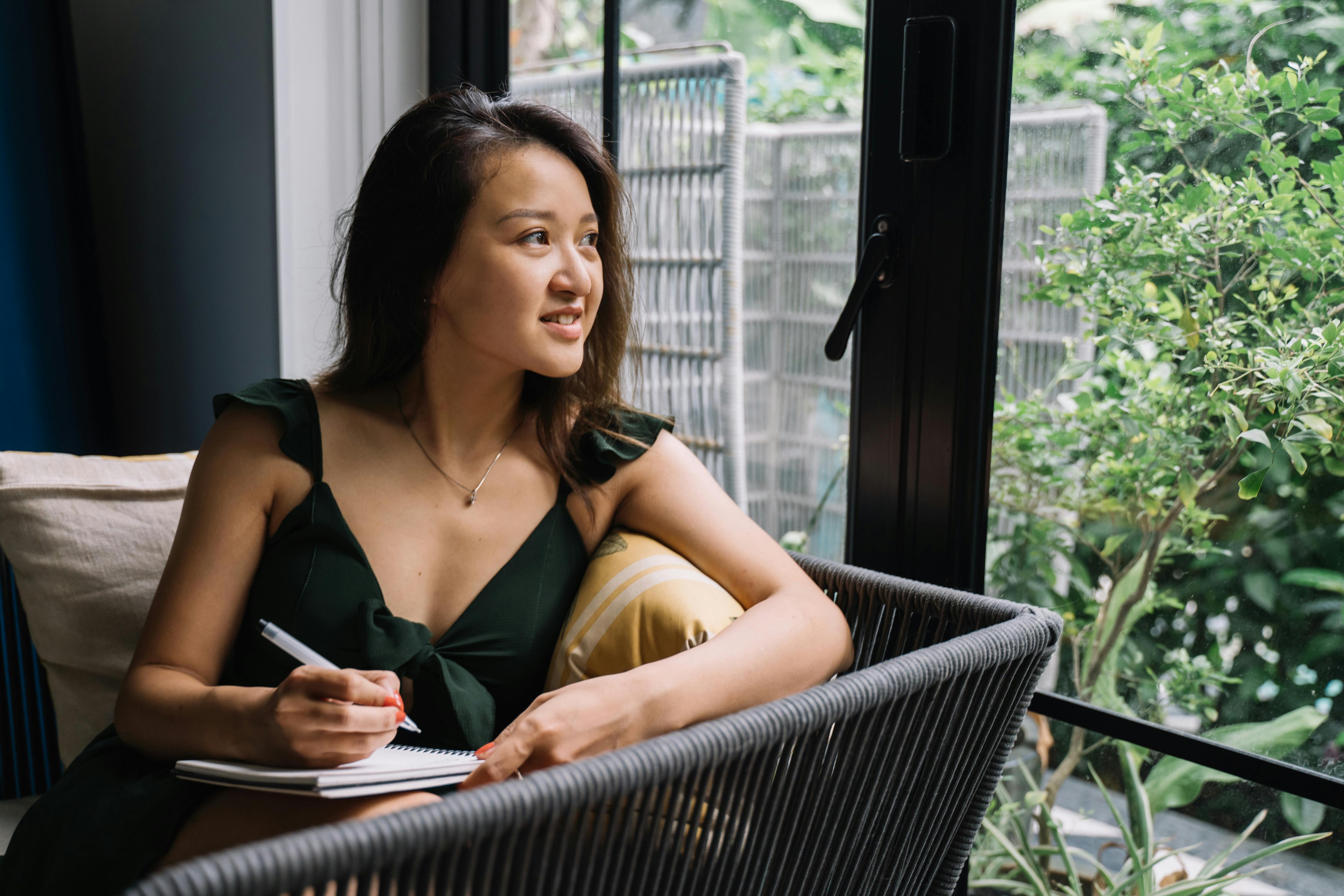 Free Asian woman in green dress writing in a notepad by the window, smiling and relaxed. Stock Photo