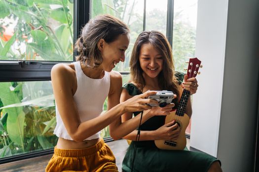 Two young women happily share a moment with an analog camera and ukulele by a window.