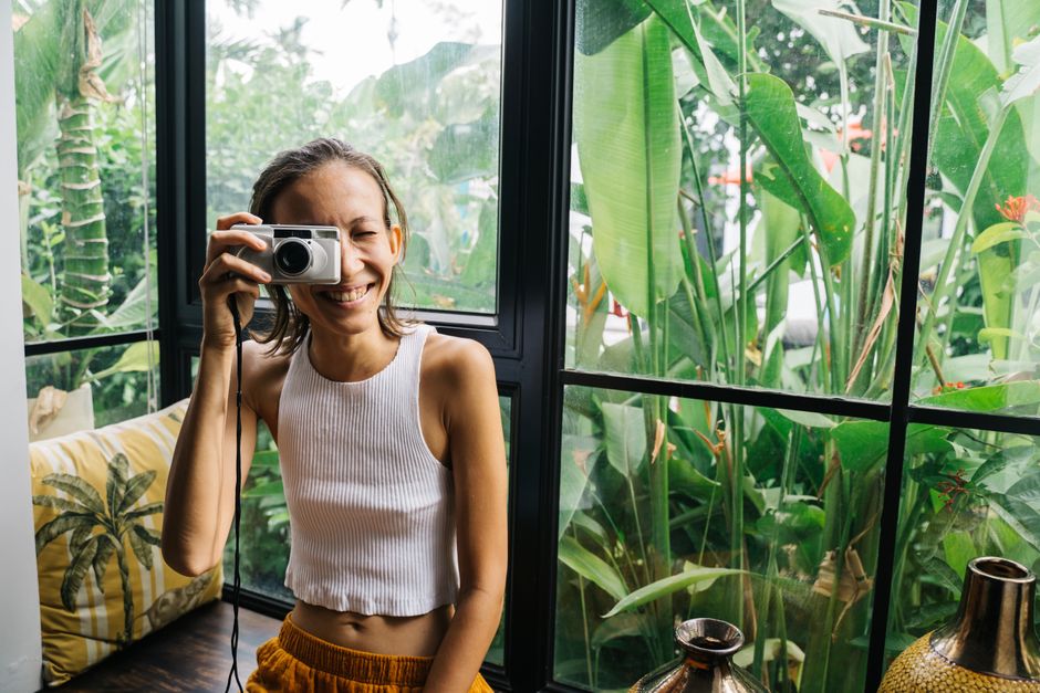 Smiling woman with a camera by a window with lush tropical plants, enjoying a fun indoor moment.