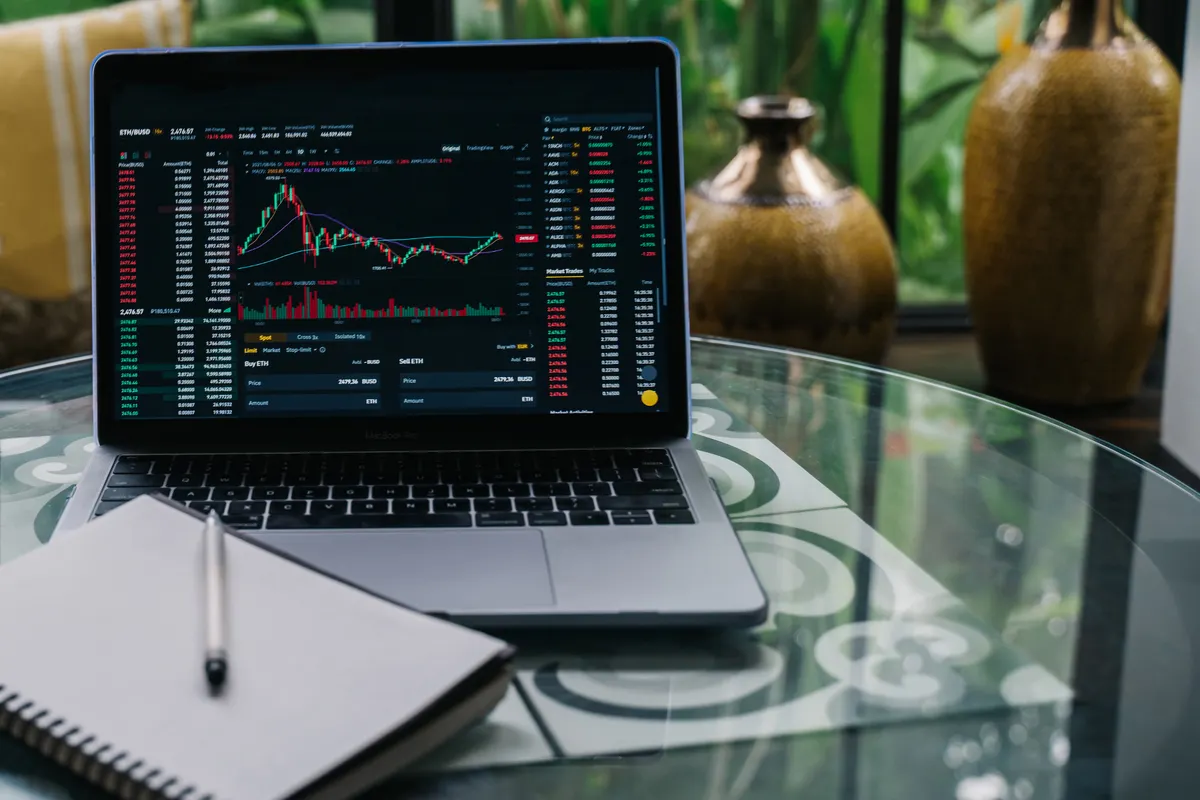 A laptop displaying cryptocurrency charts and graphs on a glass table beside a notepad, the research stage before committing to a bot subscription.