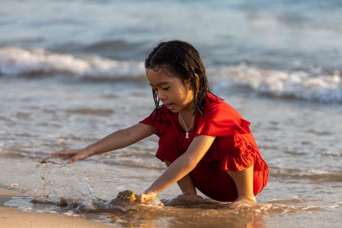 Free A child in a red dress plays joyfully at the beach by the shore on a sunny day. Stock Photo