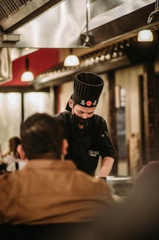 A chef wearing a black toque prepares a meal for seated diners in a restaurant.