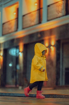 A child wearing a yellow raincoat and red boots walks alone in a dimly lit urban area on a rainy evening.