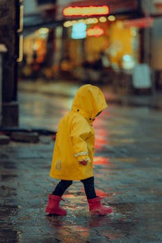 A child in a bright yellow raincoat splashes through puddles on a rainy city street, wearing red boots.