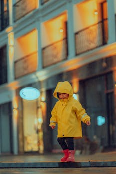 Child wearing a yellow raincoat and red boots stands on a city street under the rain at night.