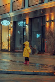 A child in a yellow raincoat stands under warm lights on a rainy evening street.