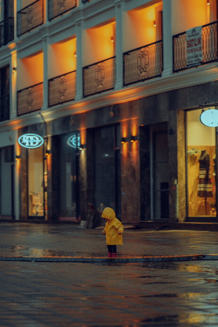 A Child In A Yellow Raincoat On A Wet Sidewalk