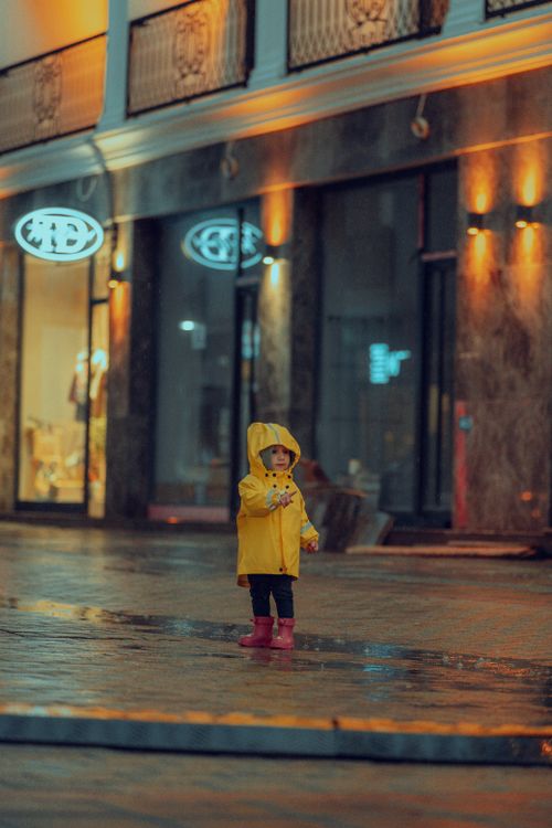 Free A young child wearing a yellow raincoat stands in a wet urban street, illuminated by warm lights at night. Stock Photo