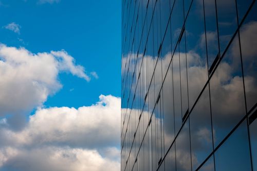 Free A modern skyscraper with reflective glass panels mirroring a blue sky and white clouds. Stock Photo