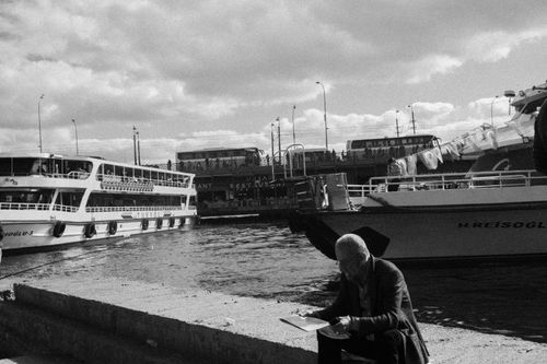 Free Monochrome scene of an elderly man sitting by a pier in İstanbul with ferries docked nearby. Stock Photo