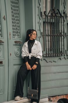 Stylish woman in black pants and white jacket standing by an ornate door, exuding sophistication.