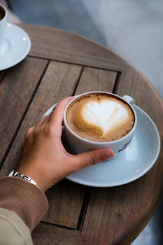 A cozy moment with a heart-patterned latte in a white cup on a wooden table, held by hand.