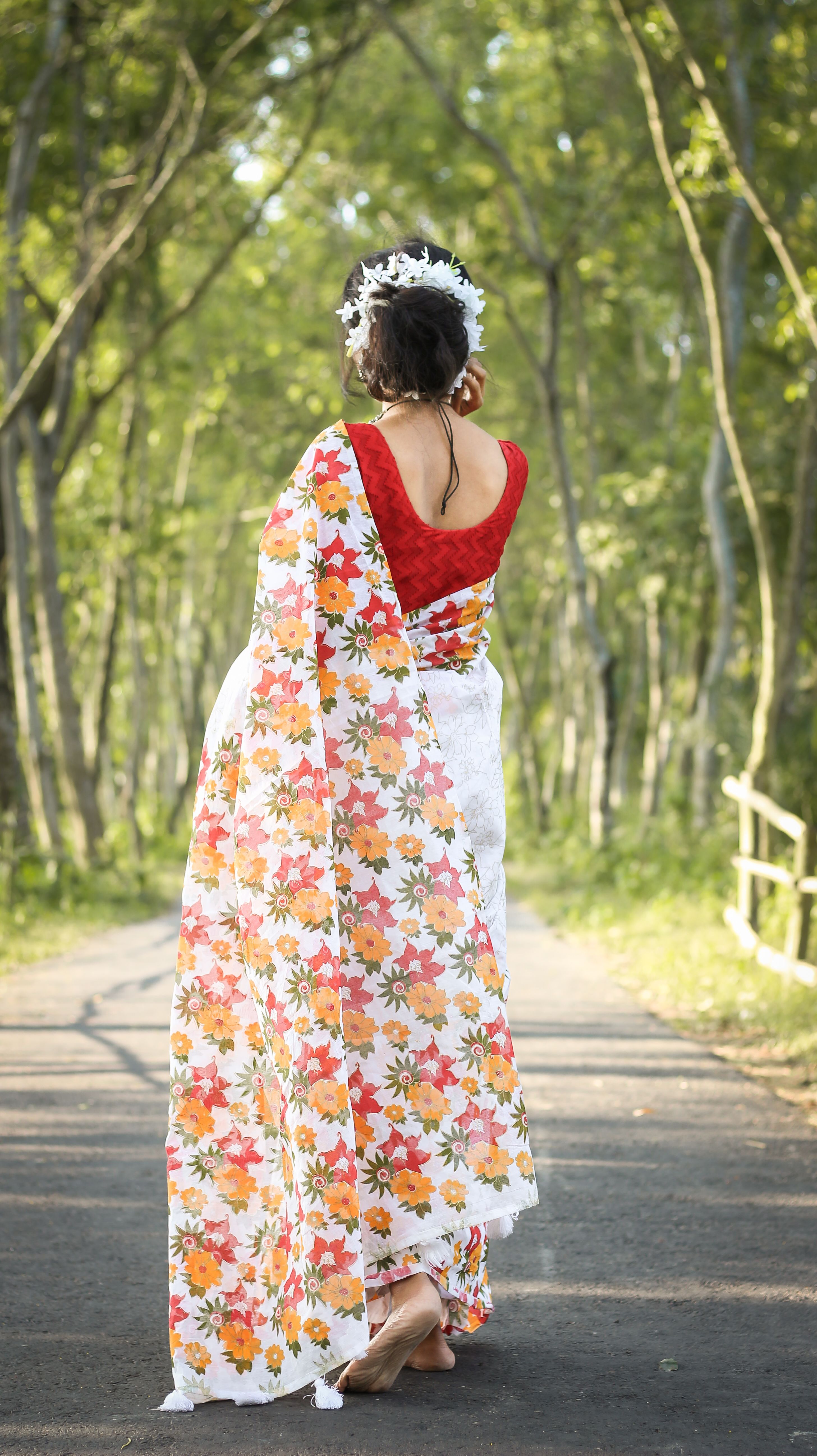 A woman in a floral sari walks barefoot on a forest pathway, enjoying a bright day outdoors.