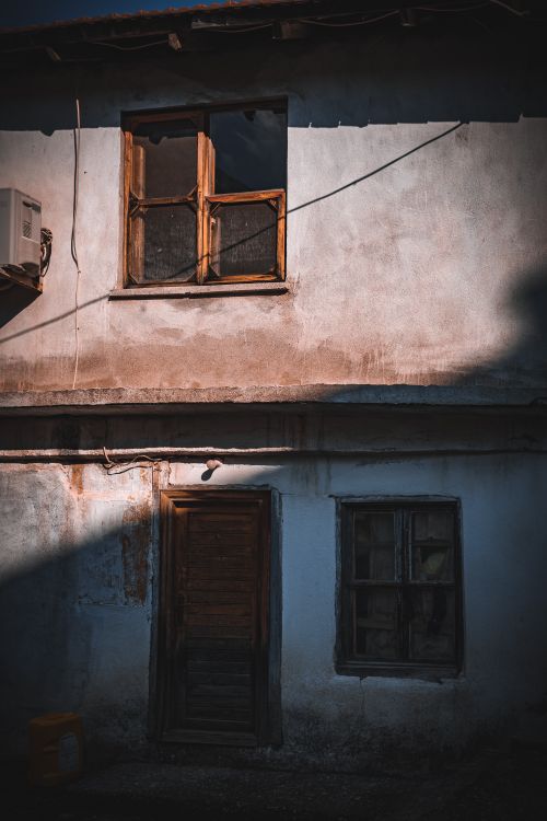 Free A shadow-cast rustic building exterior showcasing a run-down house with weathered windows and doors. Stock Photo