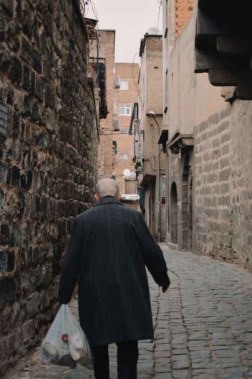 Free An elderly man with a shopping bag walks through a narrow cobblestone alleyway in an urban setting. Stock Photo