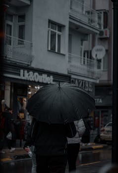 Pedestrians navigate a rainy city street, carrying black umbrellas by shops and buildings.