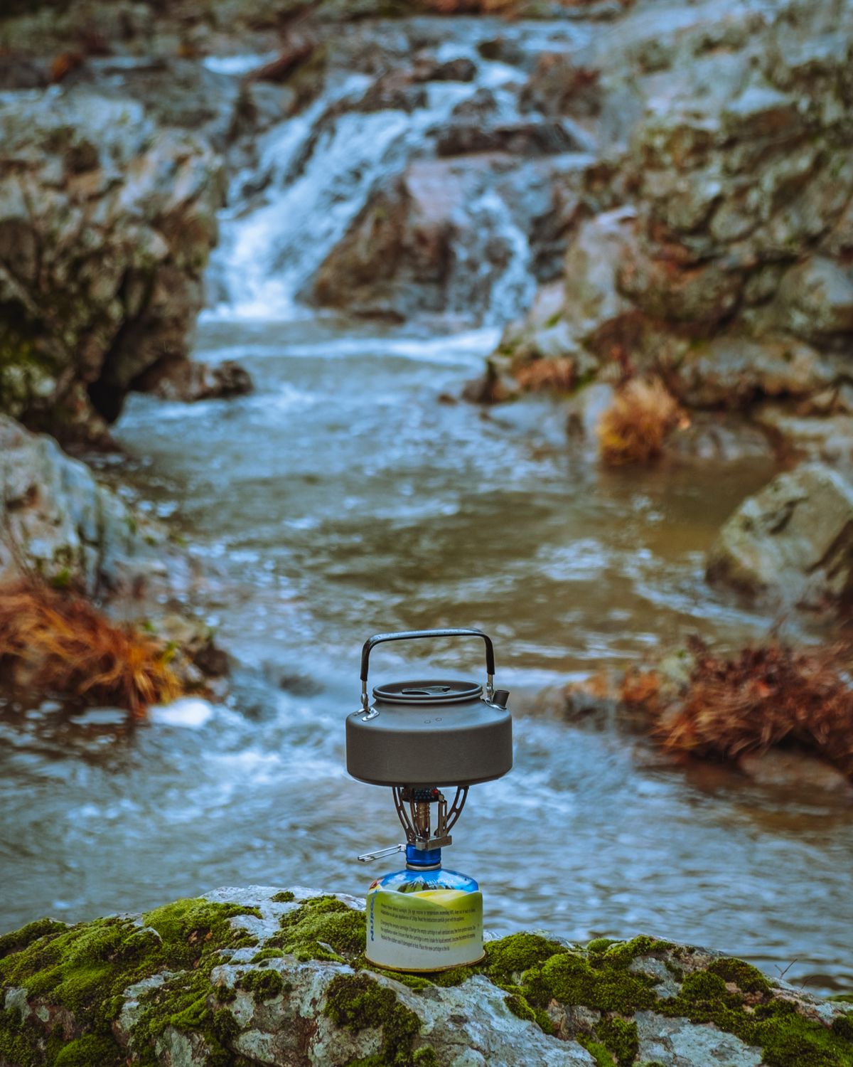 Camping kettle on a portable stove beside a rocky stream