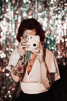 Young woman with tattoos holding an instant camera against a sequin background, capturing party moments.