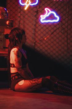 A woman with long hair and tattoos sits under neon lights, creating an urban, edgy atmosphere.