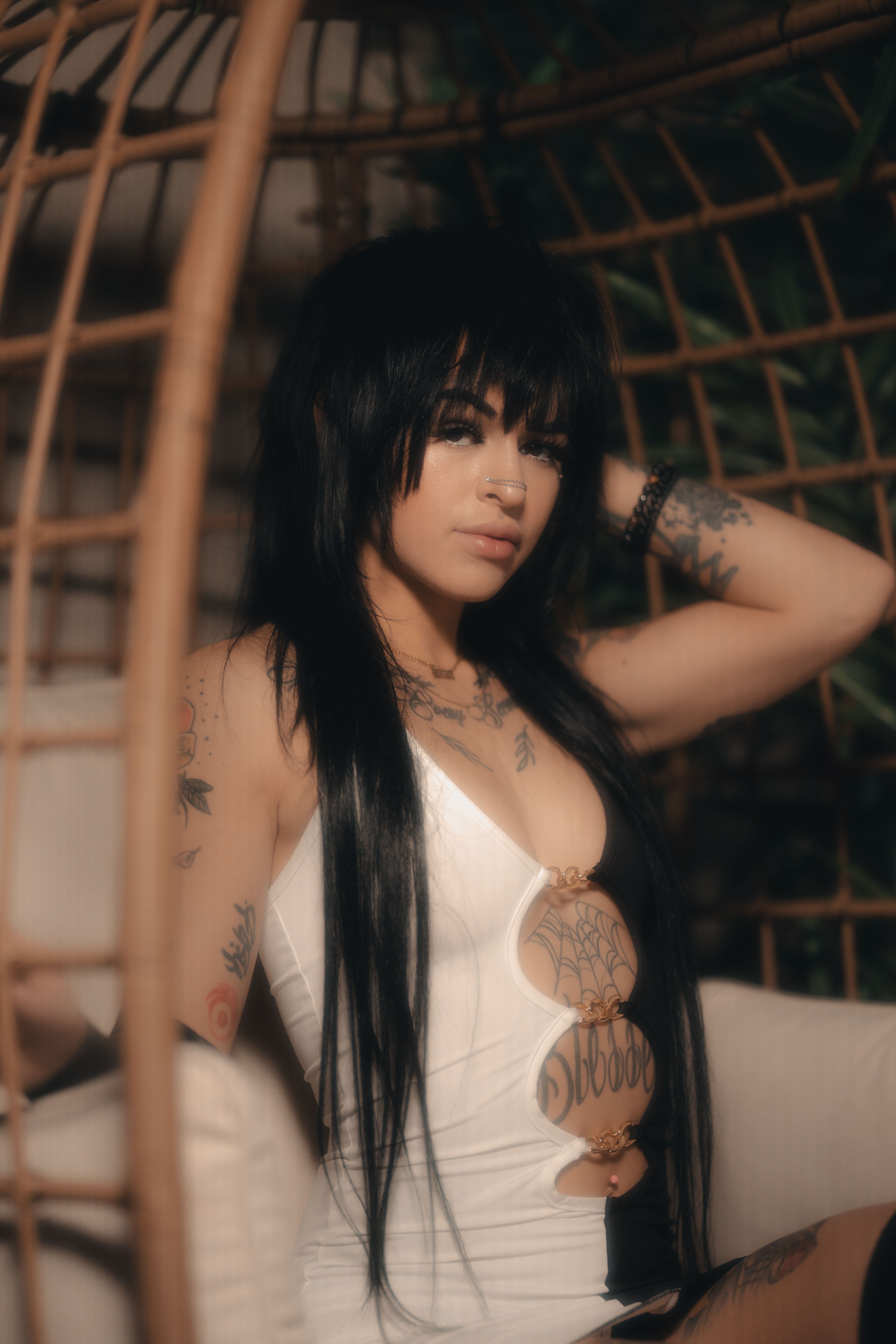 Woman with long black hair and tattoos poses stylishly in a wicker chair indoors.