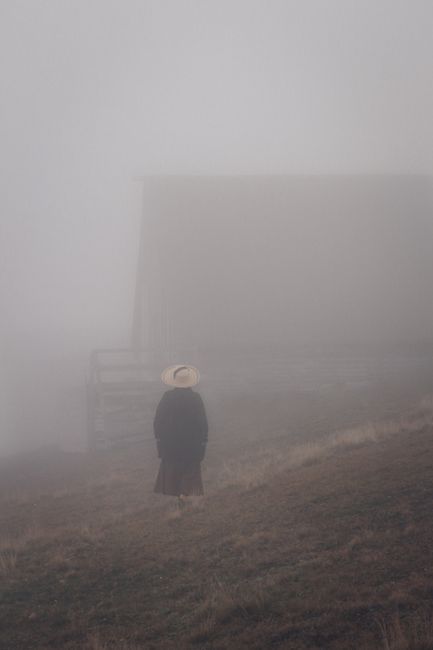 A solitary figure in a hat stands in a foggy rural area, creating a moody atmosphere.