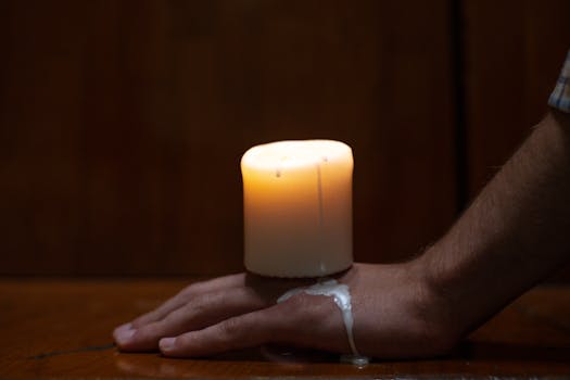Hand with lit candle drips wax in a dark, intimate setting on a wood surface.