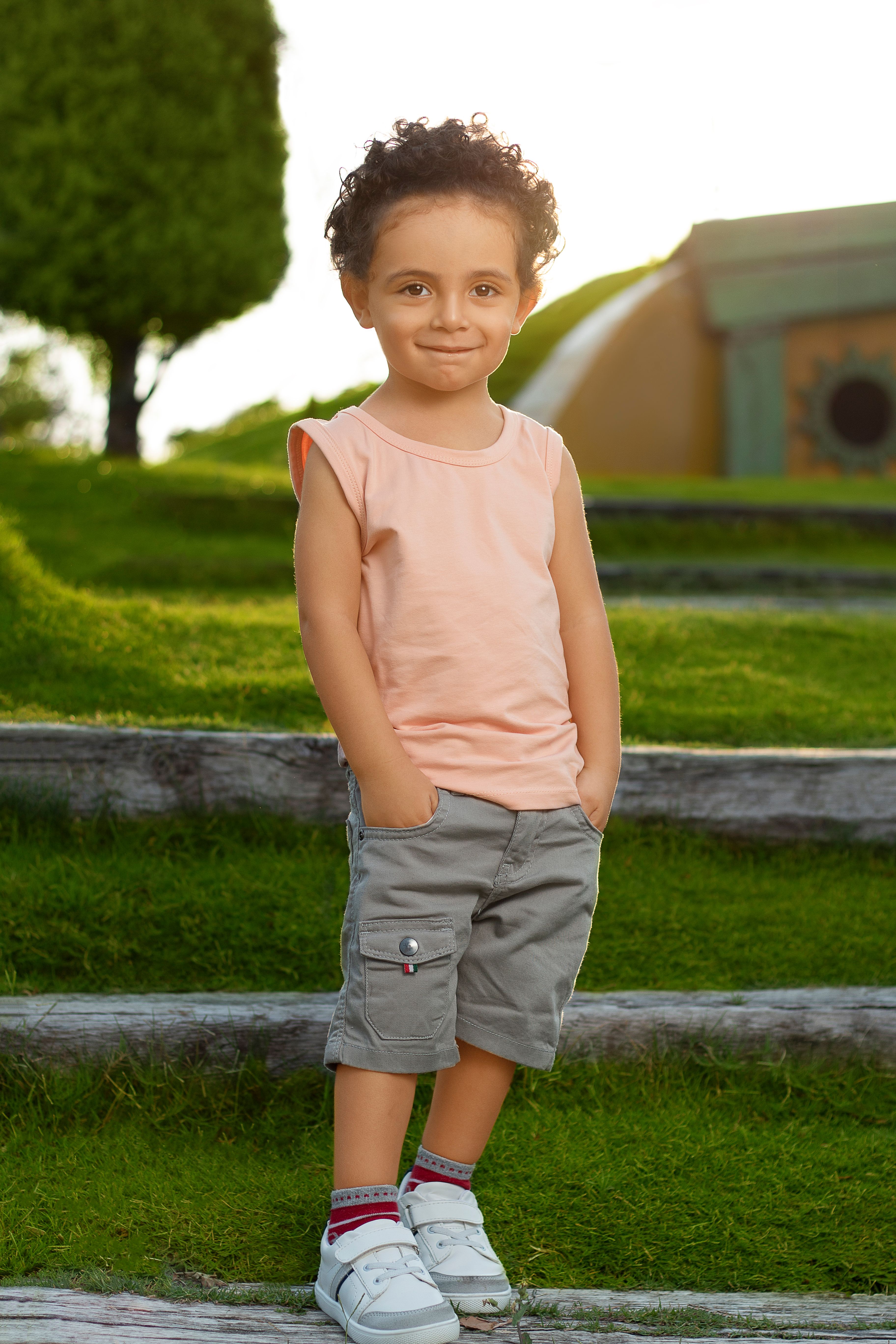 A young boy standing outdoors, smiling in a trendy summer outfit with hands in pockets.