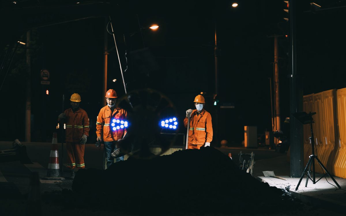 Workers in reflective gear and helmets conduct roadworks at night under bright lights.
