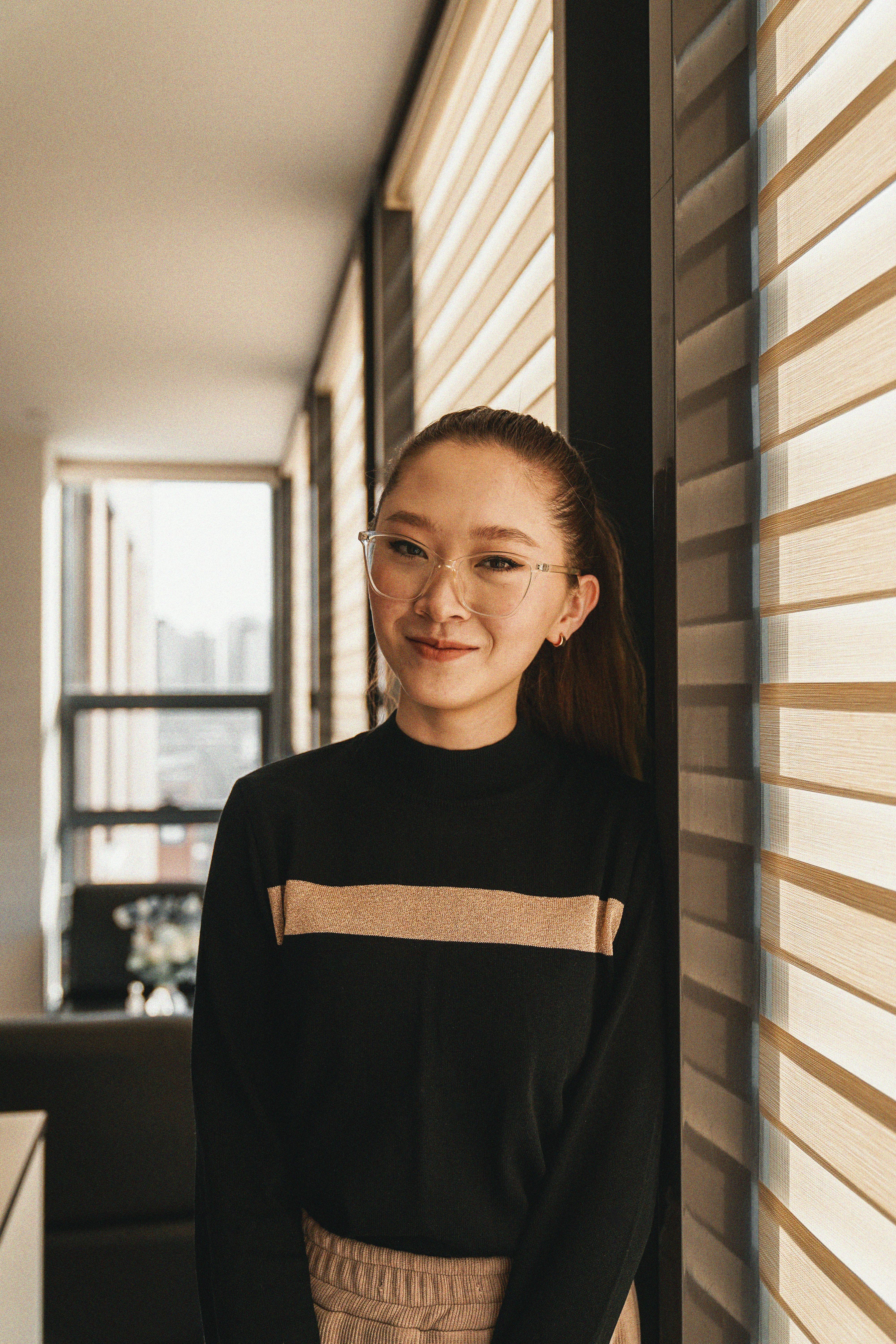 young woman in eyeglasses standing next to a window and smiling